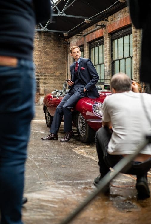 A man in a suit having his photo taken leaning on a jaguar sports car