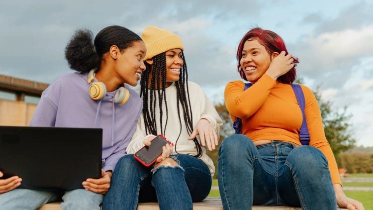 Three multiracial teenage girls chatting happily and using technology outside the university campus